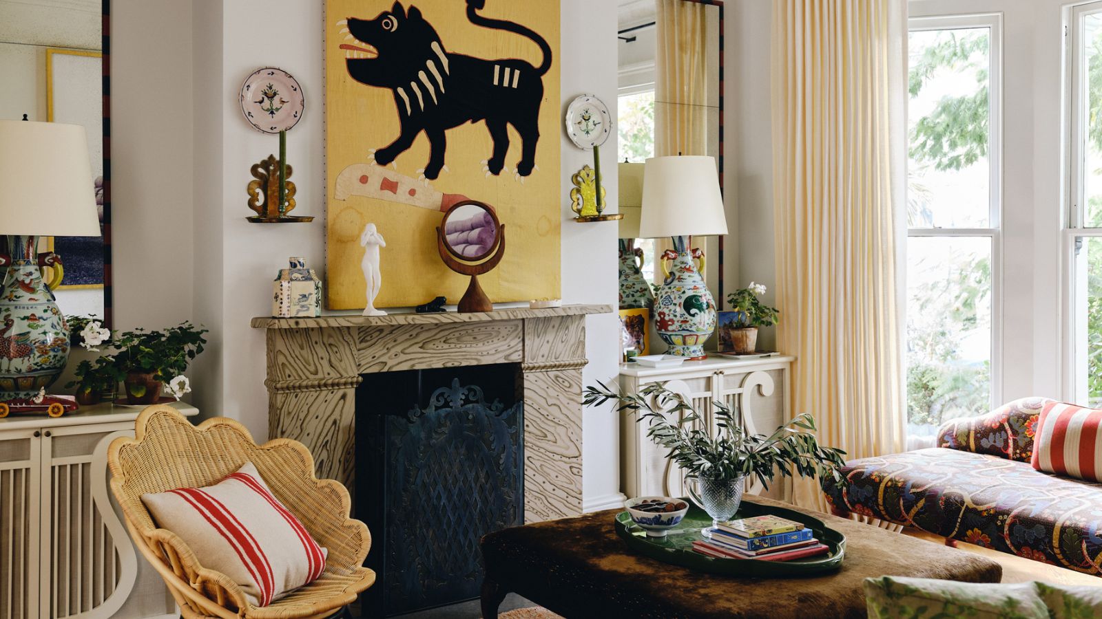image of an eclectic living room with white walls and a large yellow painting above a marble fireplace with a scalloped rattan chair and eclectic furniture