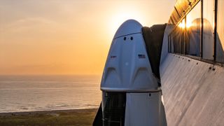 The crew access arm at spacex's launch tower rests against the side of a white space capsule. The ocean and sunrise are in the background.