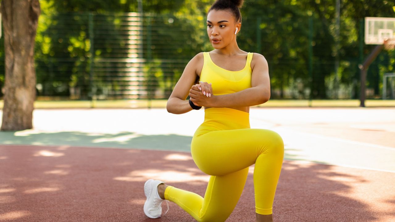 mixed race woman doing a lunge with a twist facing the camera wearing a bright yellow crop top and leggings and white trainers in a park with trees behind her 