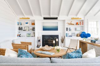 beach house living room with white walls and ceiling and pale blue sofa and rattan chairs grouped round fireplace with woodburning stove and round coffee table with blue accessories and rug