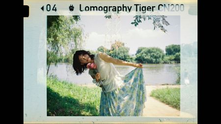 Photo of a woman in a sleeveless top and floral skirt holding flowers, posing by a lake with trees and a radio tower in the background