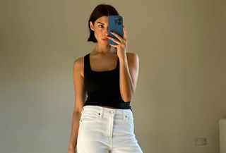 Influencer wearing black tank top and white jeans taking mirror selfie of full outfit.