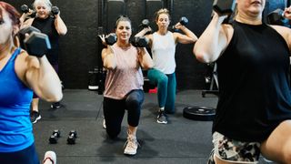 Group of women strength training in a gym class by holding dumbells at shoulder height while doing a lunge, wearing active clothes
