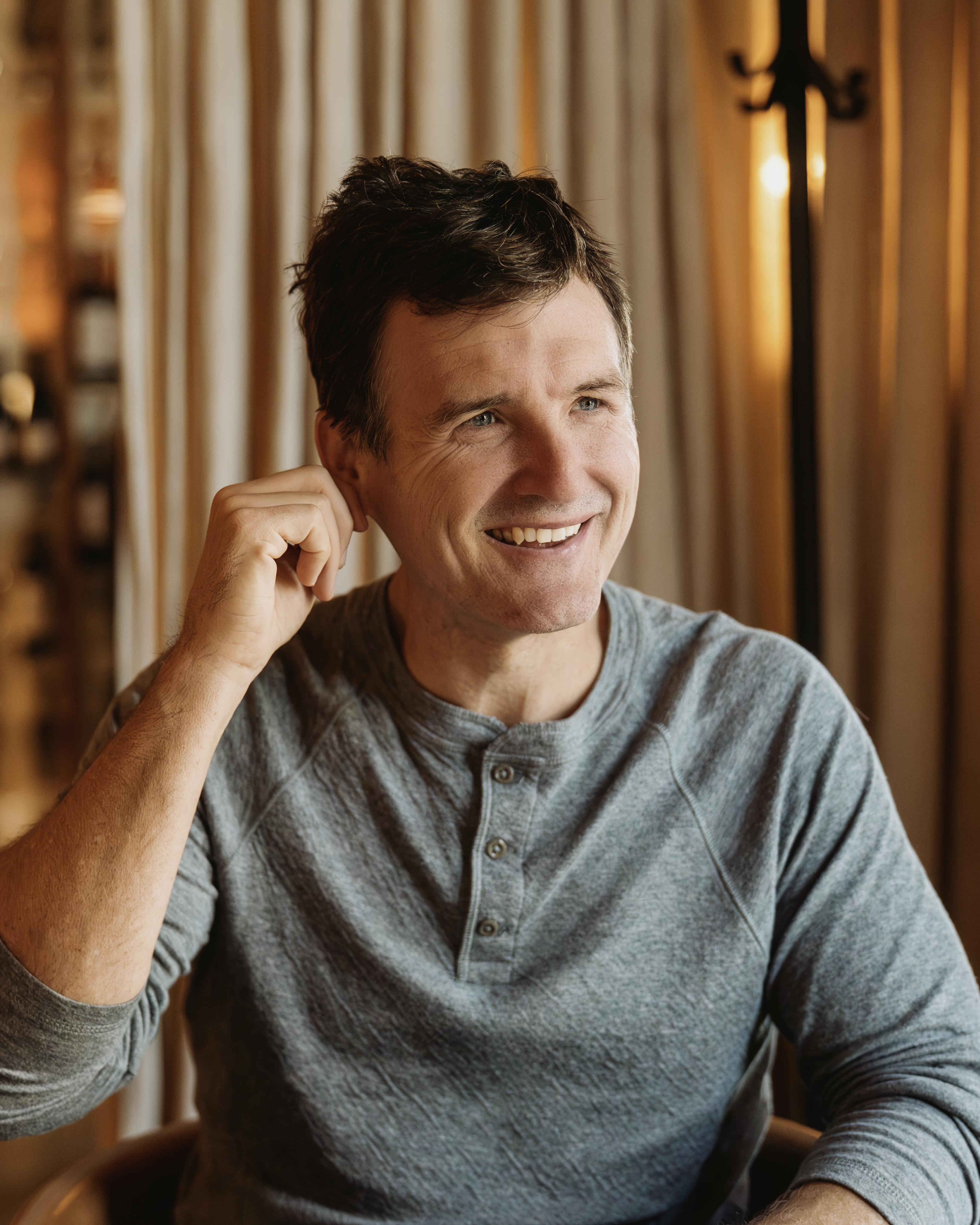 A man with brown hair wearing a grey top smiling at the camera