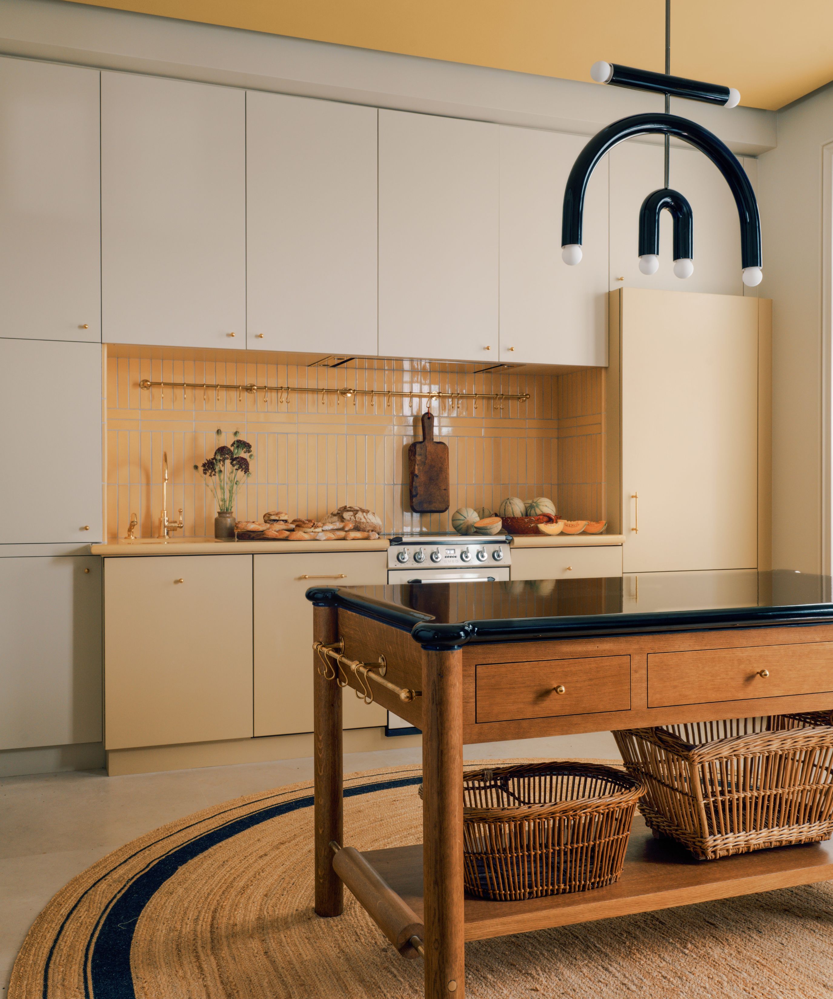 A modern minimalist kitchen with cream-colored cabinetry and a yellow-tiled backsplash. A wooden kitchen island with a black countertop sits on a circular jute rug, illuminated by a unique black sculptural pendant light.