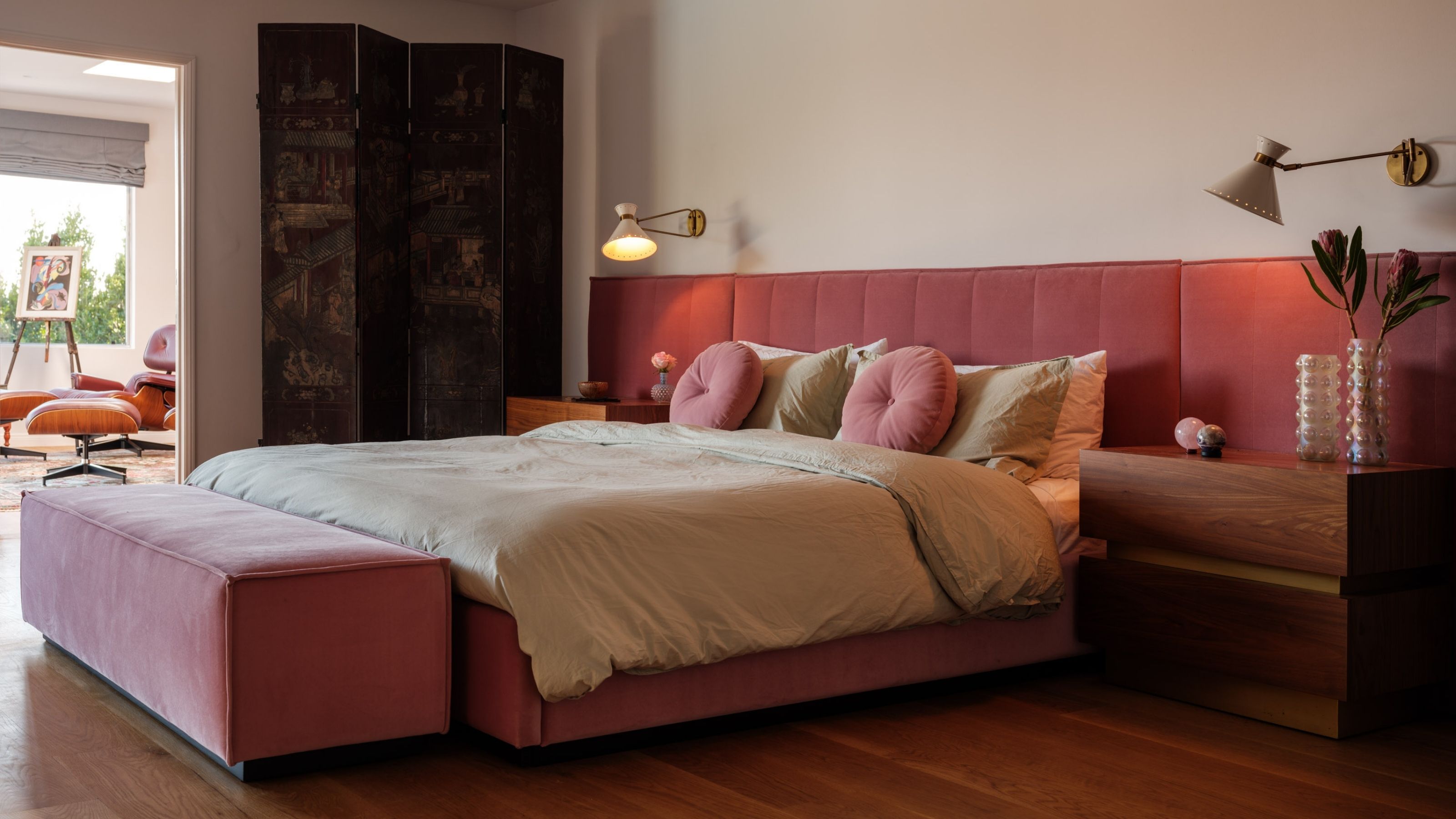 Image of a large pink bed with an extended headboard in a white bedroom with wooden floors. The bedding is pale green and there are pink and green pillows on the bed.