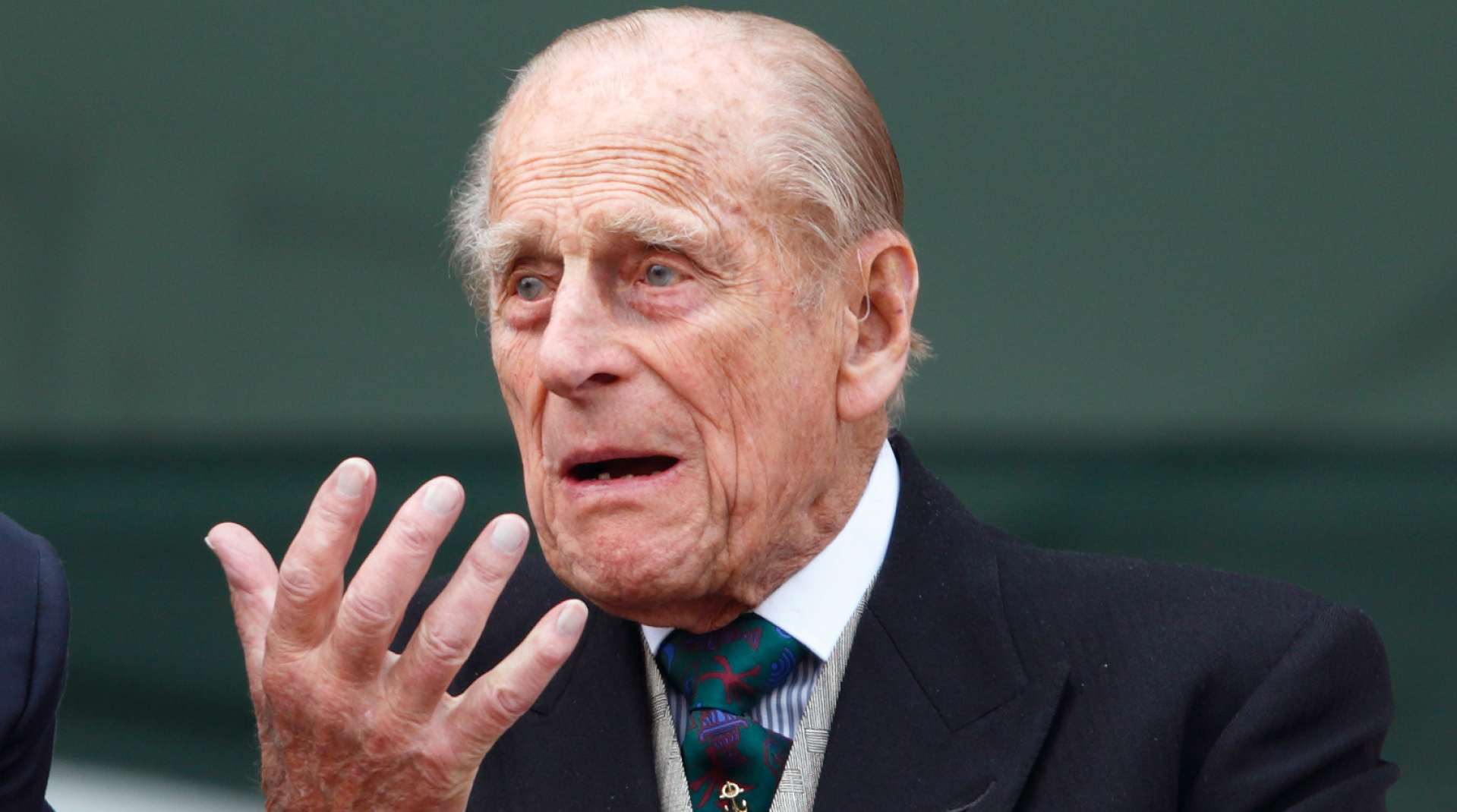 Prince Philip wears a suit with a waistcoat and tie and looks bemused while holding one hand out, he has gray hair and thinning gray hair in 2016
