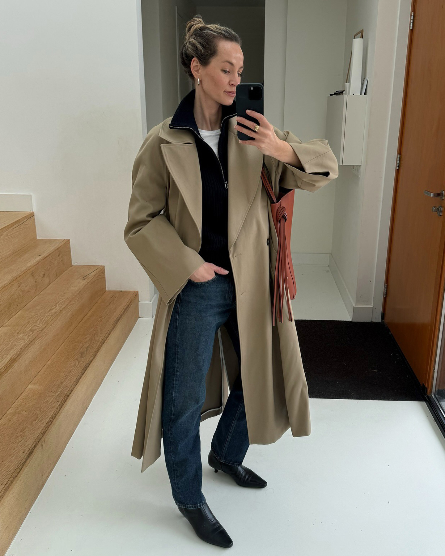 style influencer Anouk Yve poses for a mirror selfie wearing a chic fall and winter outfit with a trench coat, black half-zip sweater, dark-wash jeans, and the kitten-heel boots trend for 2025