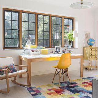 a living room with large windows with wood and black framing, a white and brown wood desk underneath the window with matching rocking chair, a yellow and wood desk chair and a rug with multi-coloured squares