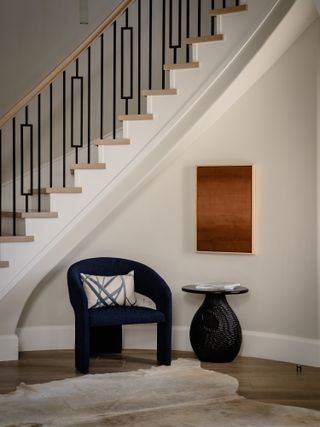 White entryway with oak and steel staircase, dark blue velvet sculptural armchair and small rounded side table with art hanging above