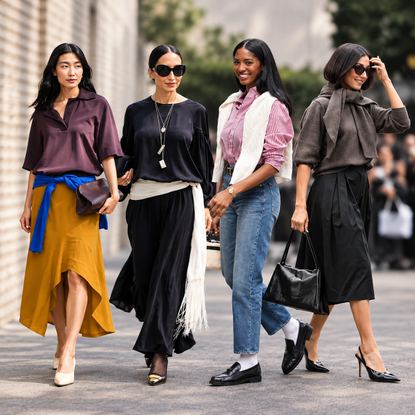 Four women in layered fall outfits, including sweaters, skirts, jeans, scarves, and loafers, walk along a city street.