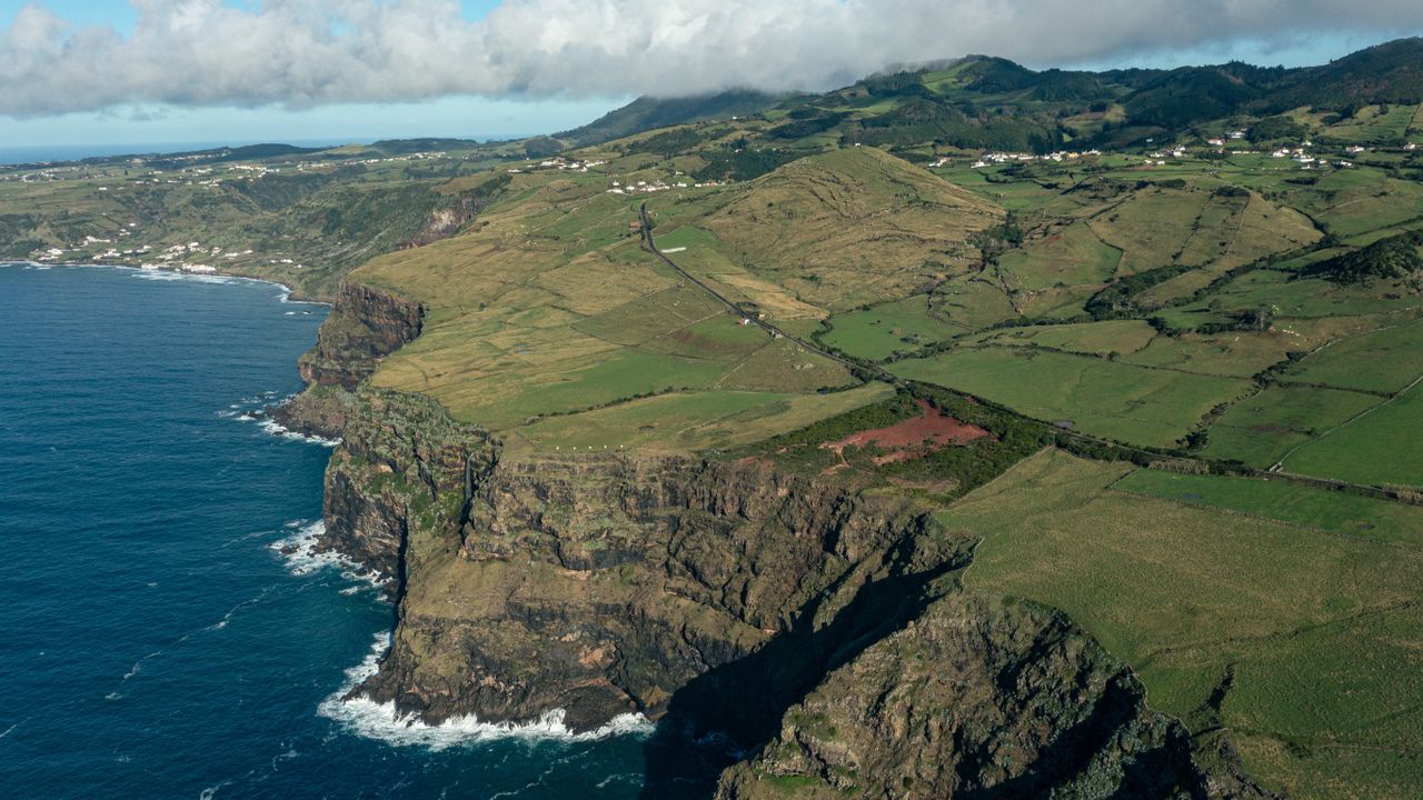 a blue ocean crashes against the bottom of a high cliff that gives way to green fields stretching to mountains in the distance.