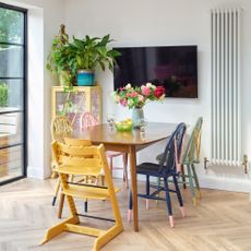 a wooden dining table with colourful upcycled dining chairs, herringbone wooden flooring, a wall-hung tv, and a tall white radiator