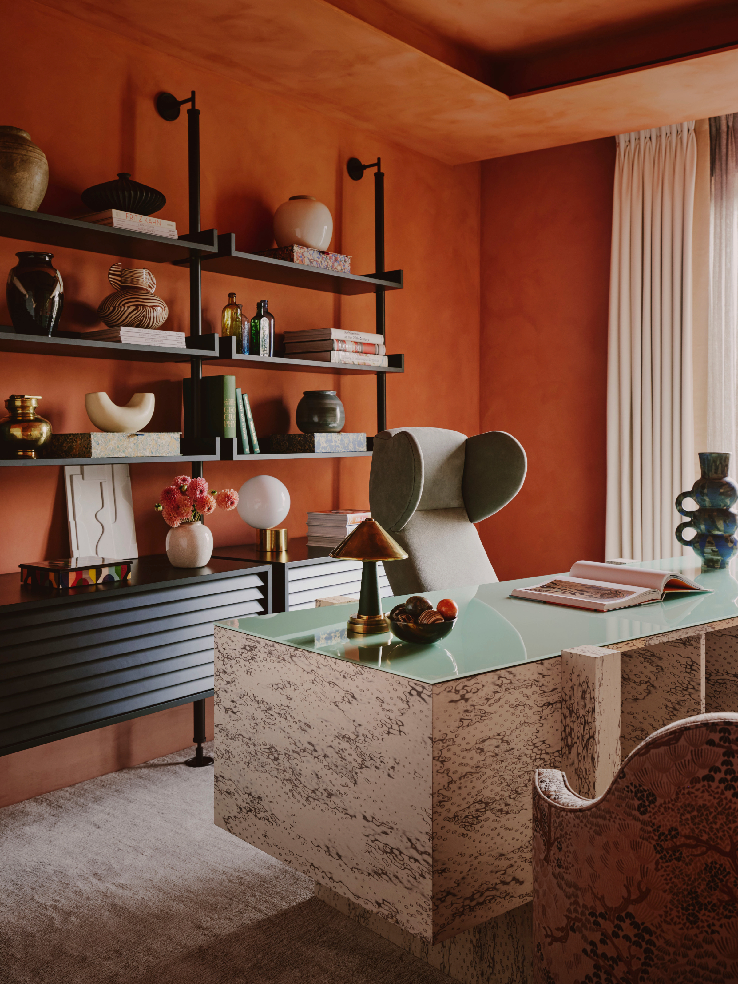 Home office with rust orange limewash effect walls, black open shelving, stone desk and grey chair