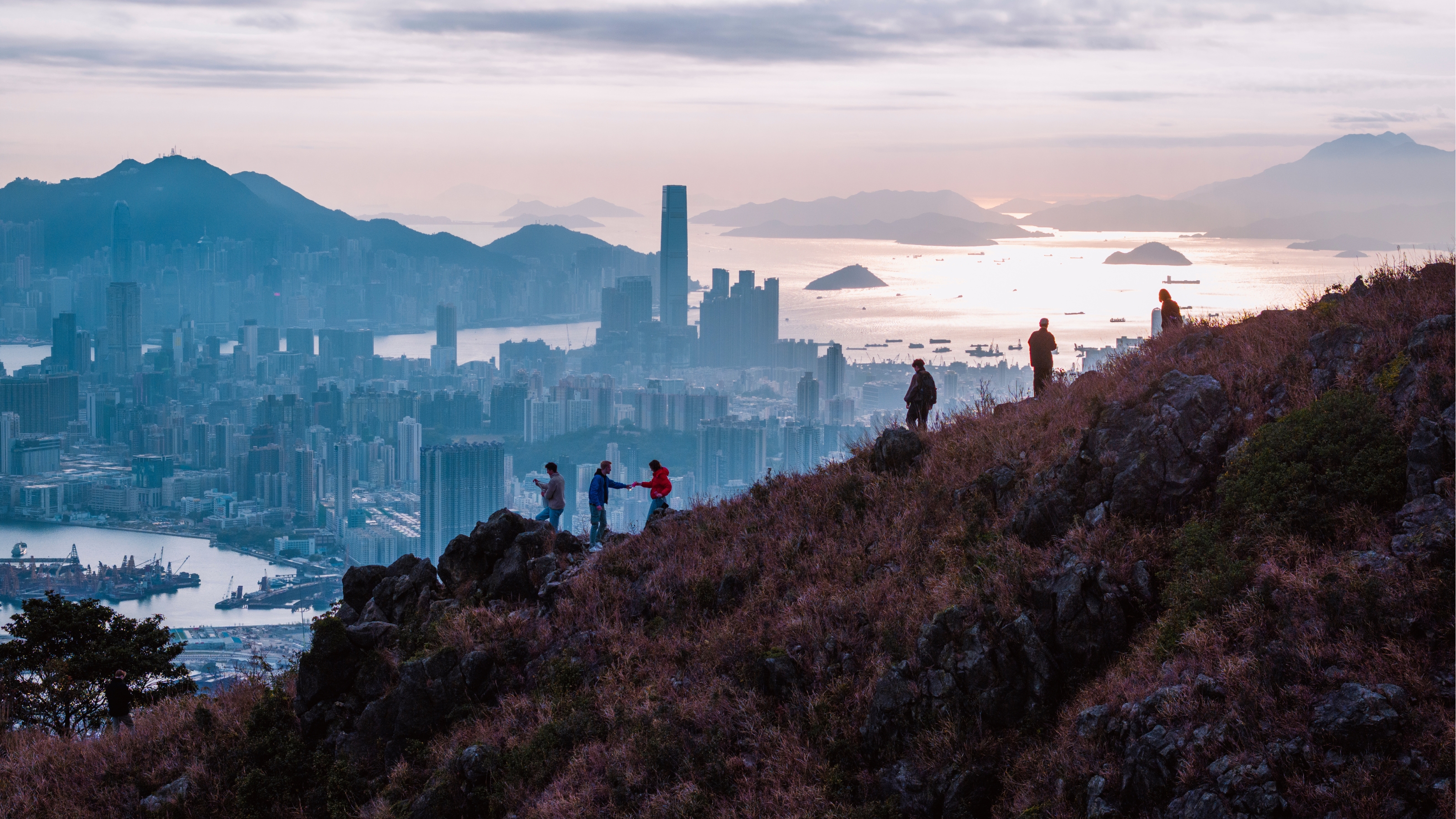 A panoramic view of a mountain slope explored by six people on foot beyond which opens up the skyline of a city filled with skyscrapers and water.