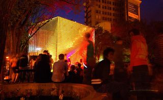 Large metal textured cube in a public square at nightime lit with bright coloured lights