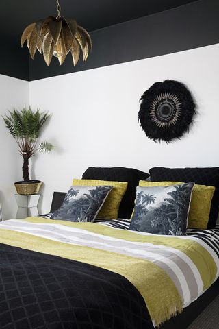 A bedroom with white wall paint and black ceiling paint decor, global-inspired palm print cushions, yellow, black and grey bedding and decorative black wall hanging