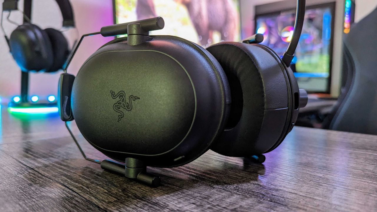 Black gaming headphones with a sleek design rest on a wooden desk. A colorful gaming setup with monitors and LED lights is visible in the background.