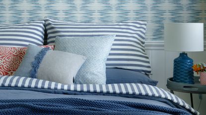 Close up of a bed stacked with blue and white striped bedding next to a small side table with a blue ceramic table lamp