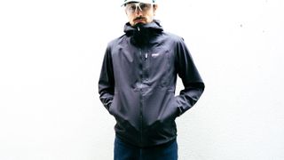 A white man in a black waterproof cycling jacket stands against a white wall