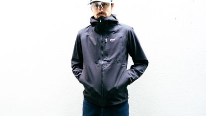 A white man in a black waterproof cycling jacket stands against a white wall