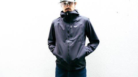 A white man in a black waterproof cycling jacket stands against a white wall