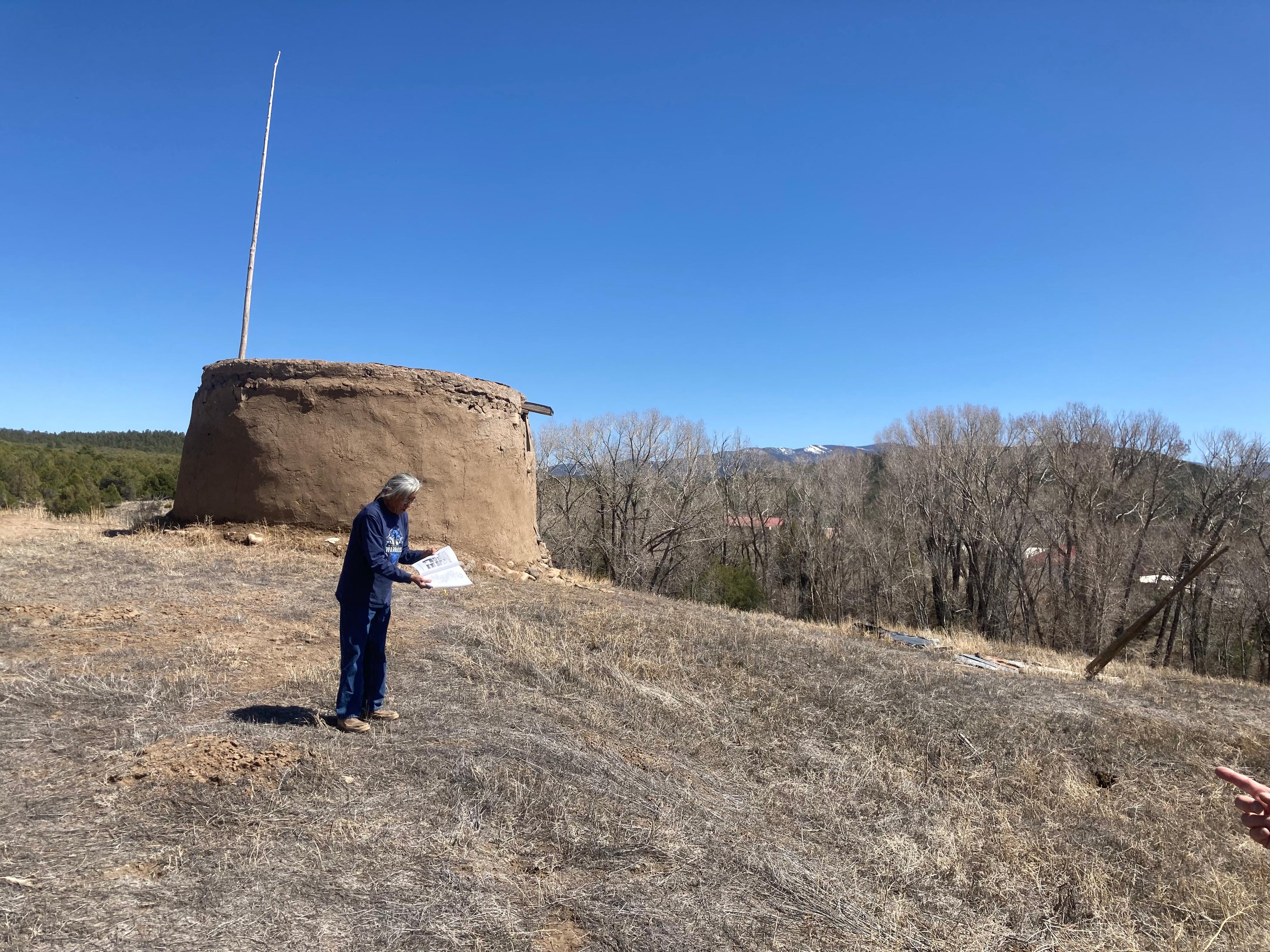 A person stands by a Picturis Pueblo round house with papers in their hand.