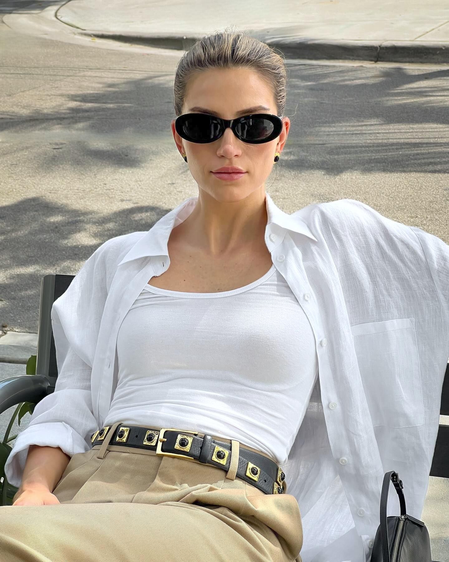 fashion influencer Cass Dimicco poses for a photo while sitting on a bench in Miami wearing black oval sunglasses, a white tank top, open white button-down shirt with pocket, studded balck Aureum belt, tank pants, and a mini black bag from The Row