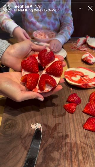 Meghan Markle holding up a bagel covered in heart-shaped strawberies on a wood table with Princess Lilibet working behind her in a heart sweatshirt