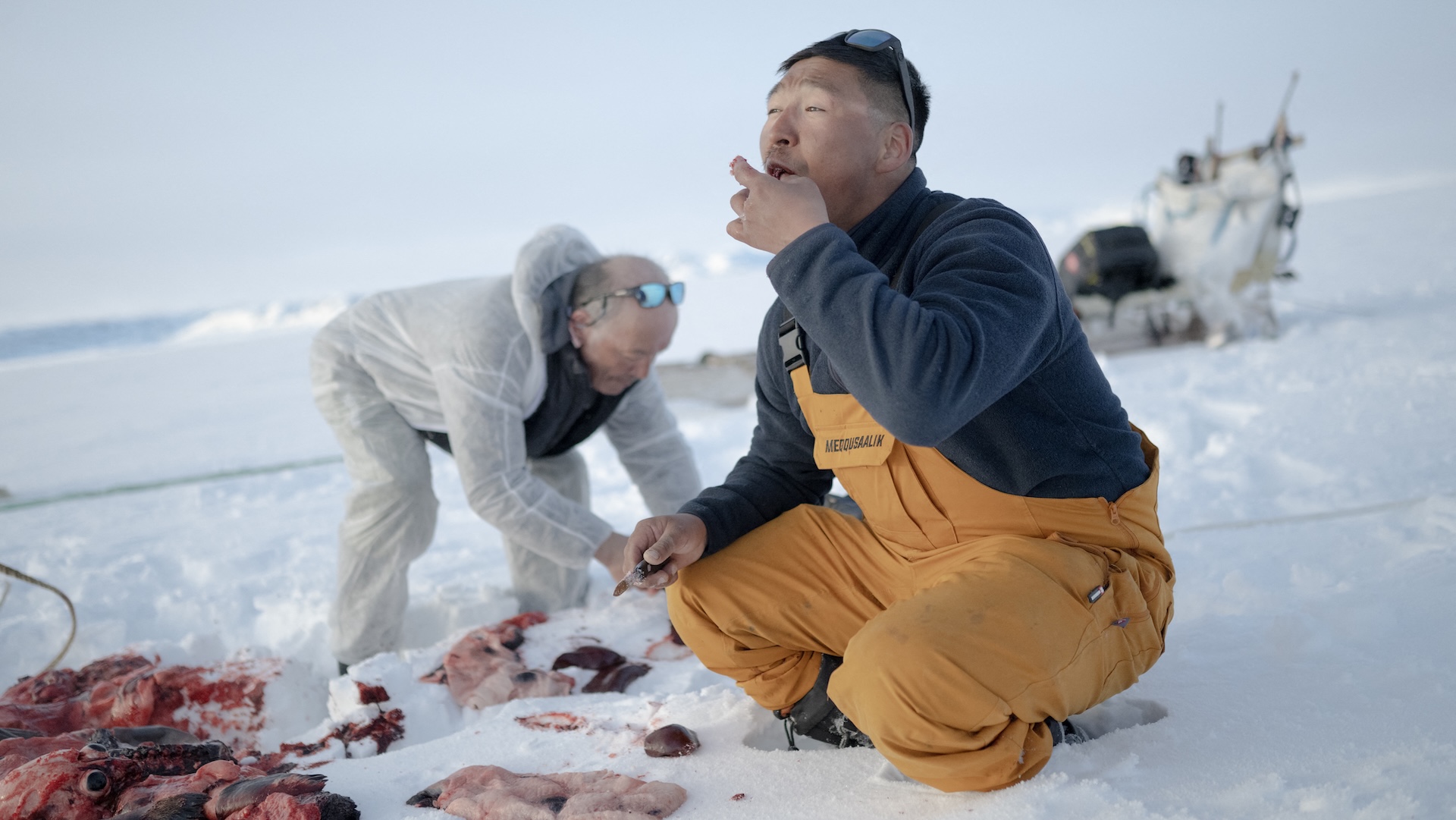 One man wearing traditional white Inuit clothes and another man wearing yellow overalls eat seal liver on the Arctic