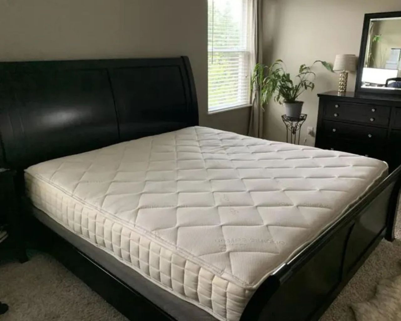 A Naturepedic Chorus Mattress on a black bedrame in a white bedroom. Beneath the bed is a grey rug, and to the right is a window with slatted blinds, a houseplant on a stand, and a matching chest of drawers to the bed with a white lamp and large mirror on top.
