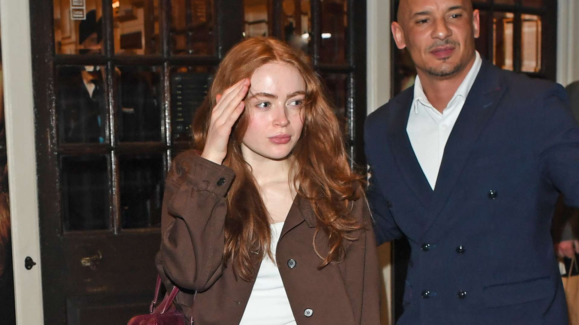 Sadie Sink wearing a Prada brown jacket, white tank top, blue bootcut jeans, burgundy bag, and black boots