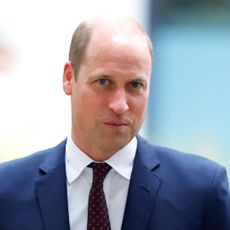 Prince William wears a blue suit with a white shirt and red tie as he smirks at the camera