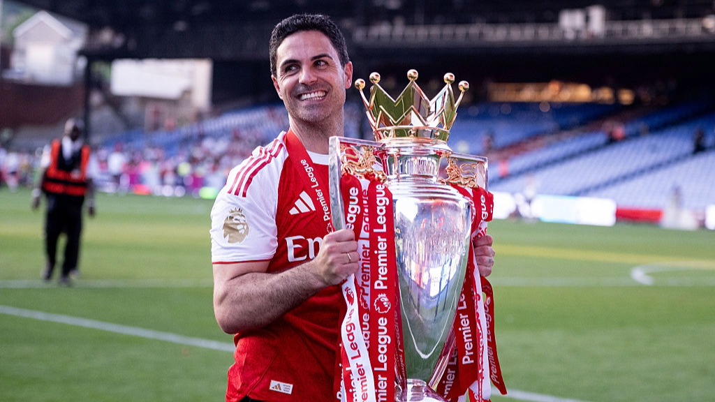 A smiling Mikel Arteta, wearing an Arsenal shirt, holds the Premier League trophy, which is decorated in red and white ribbons