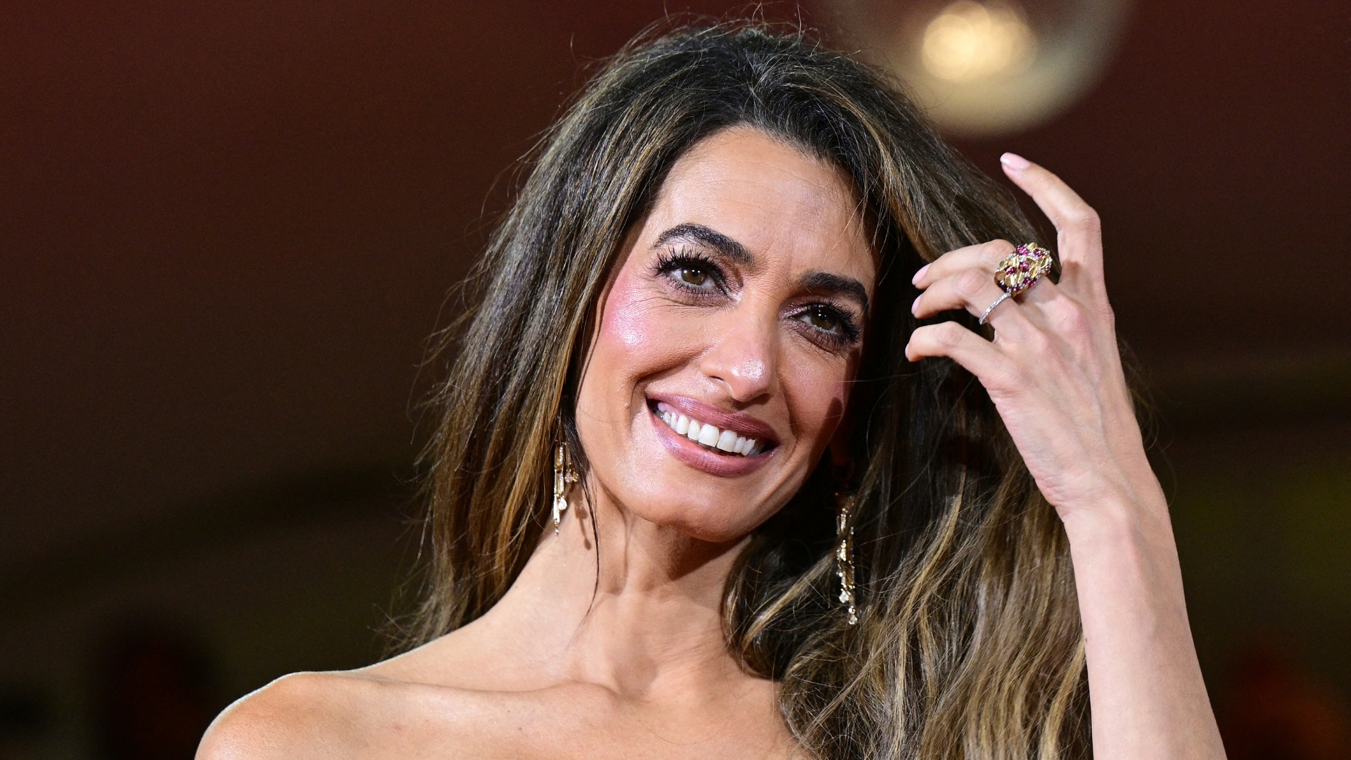 Image of Amal Clooney smiling with hand sweeping through side-part long brunette hair 