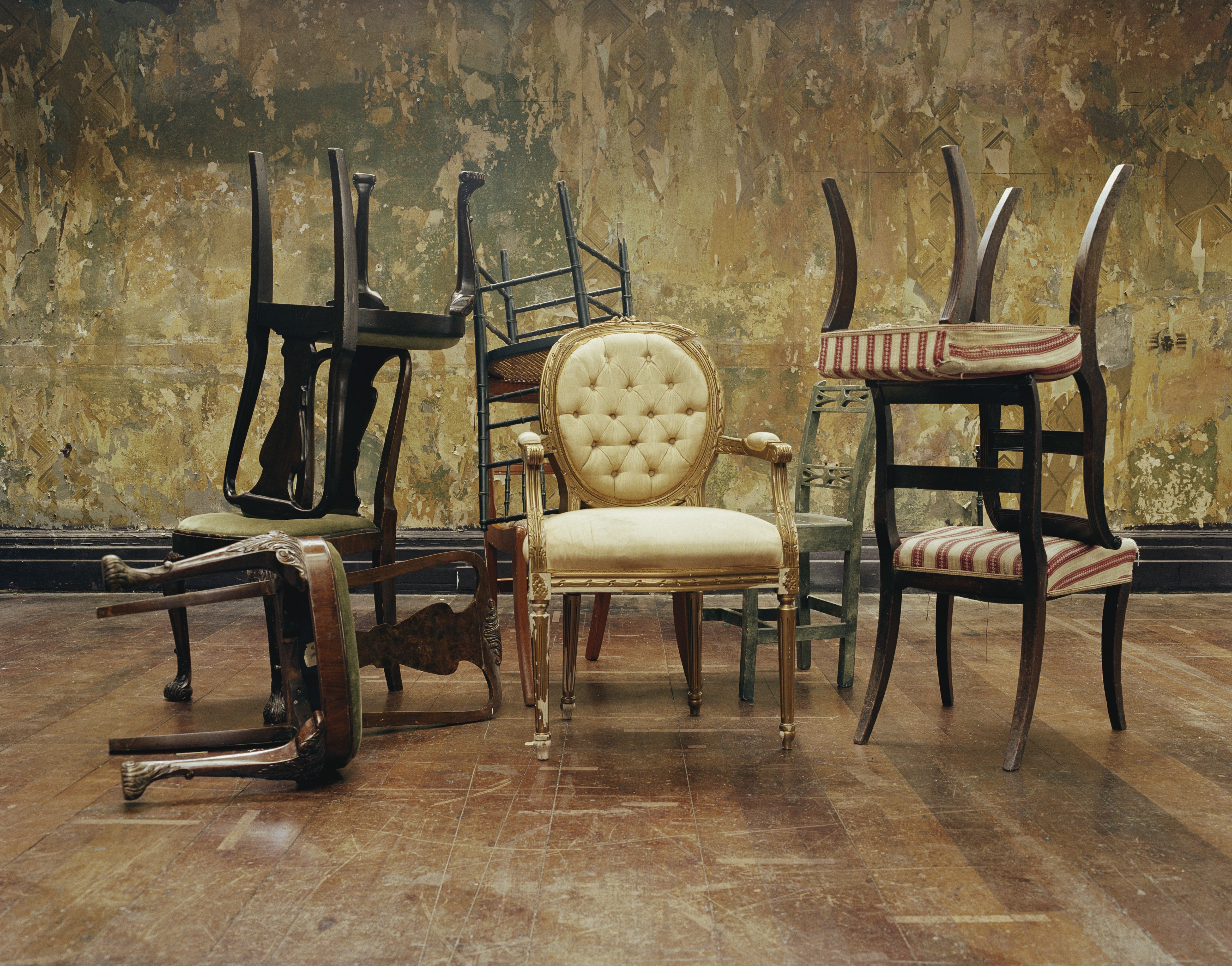 Several antique chairs arranged in a haphazard manner in a room with a dark wood floor and distressed walls