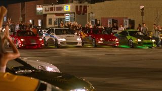 Four cars lined up for a drag race, surrounded by cars lining the street