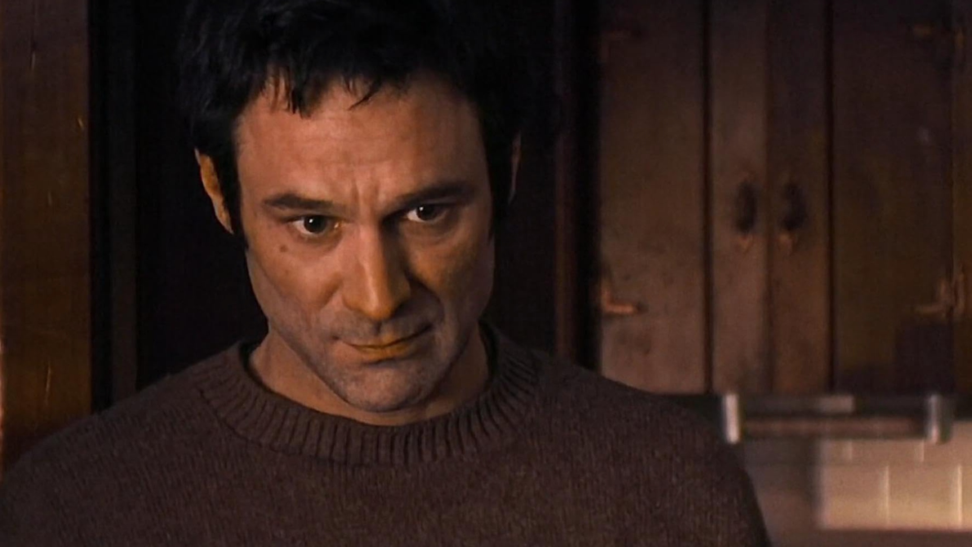 A man with short dark hair and light stubble looks onwards just past the camera in his brown sweater