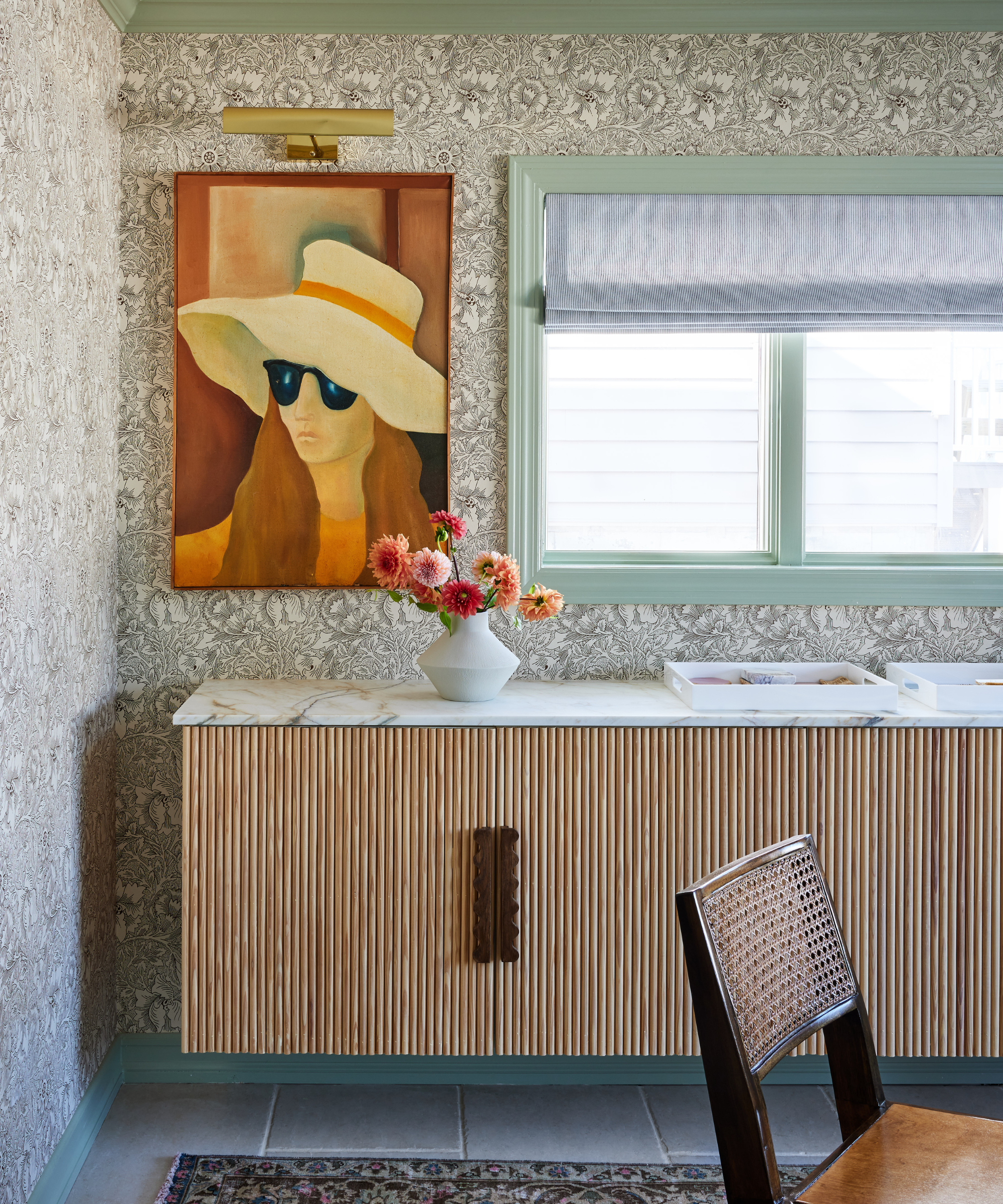 A home bar with fluted wood detailing against a wall with floral wallpaper, sage green trim, and a large painting of a woman wearing a sun hat