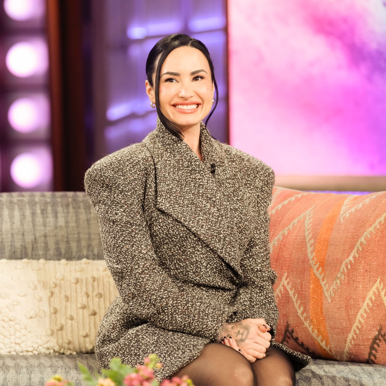 a young woman with dark hair sits on a couch on the set of a talk show