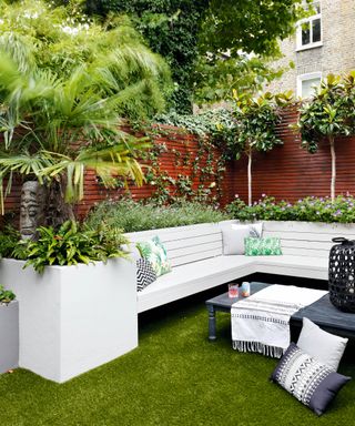 A built in outdoor corner sofa with light coloured cushions in front of a slatted fence with built in plant beds behind growing bay trees