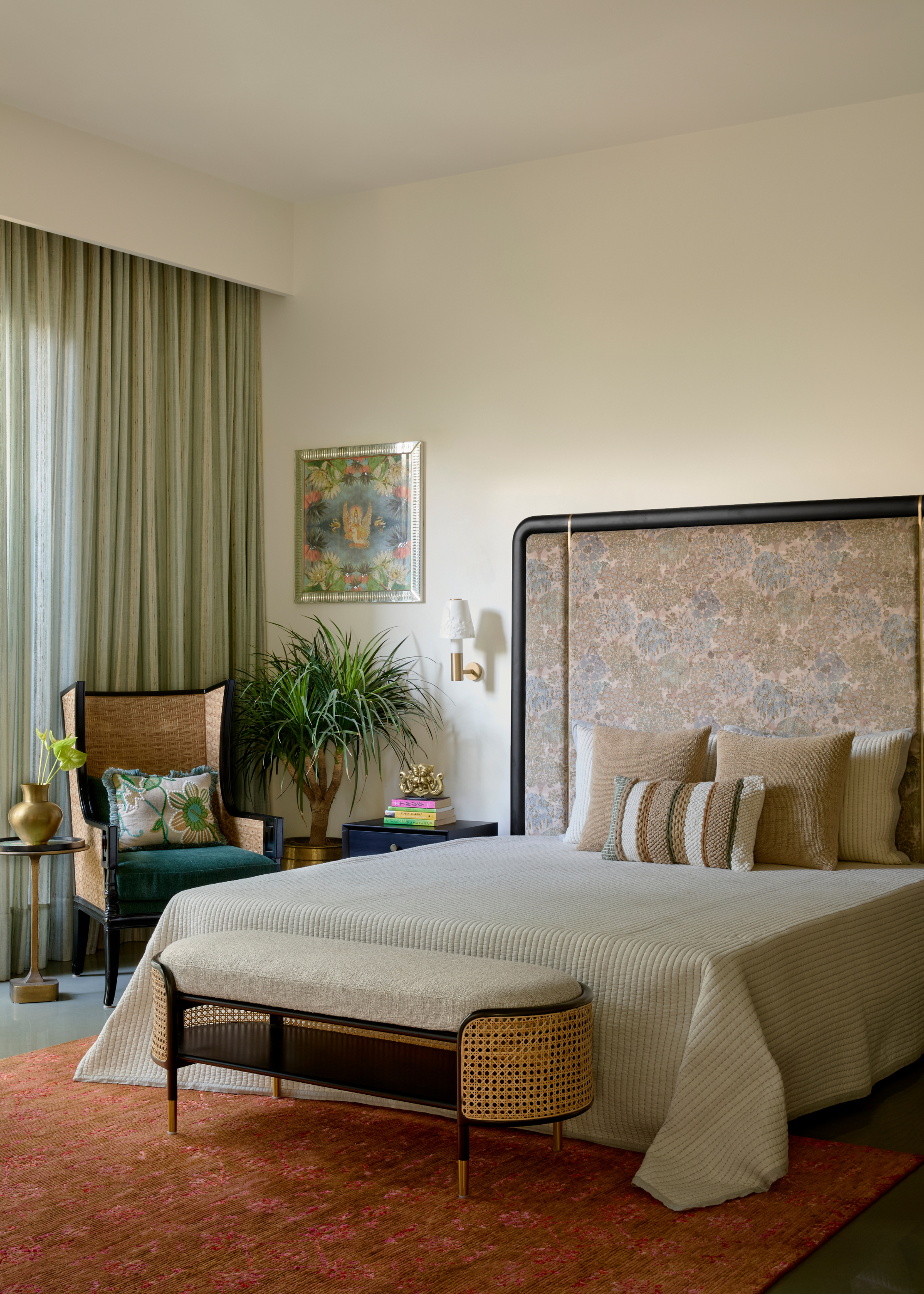 An off-white bedroom with an oversized headboard, a set of beige textured pillows, a rattan style bench, and a potted plant in a gold planter by a rattan chair