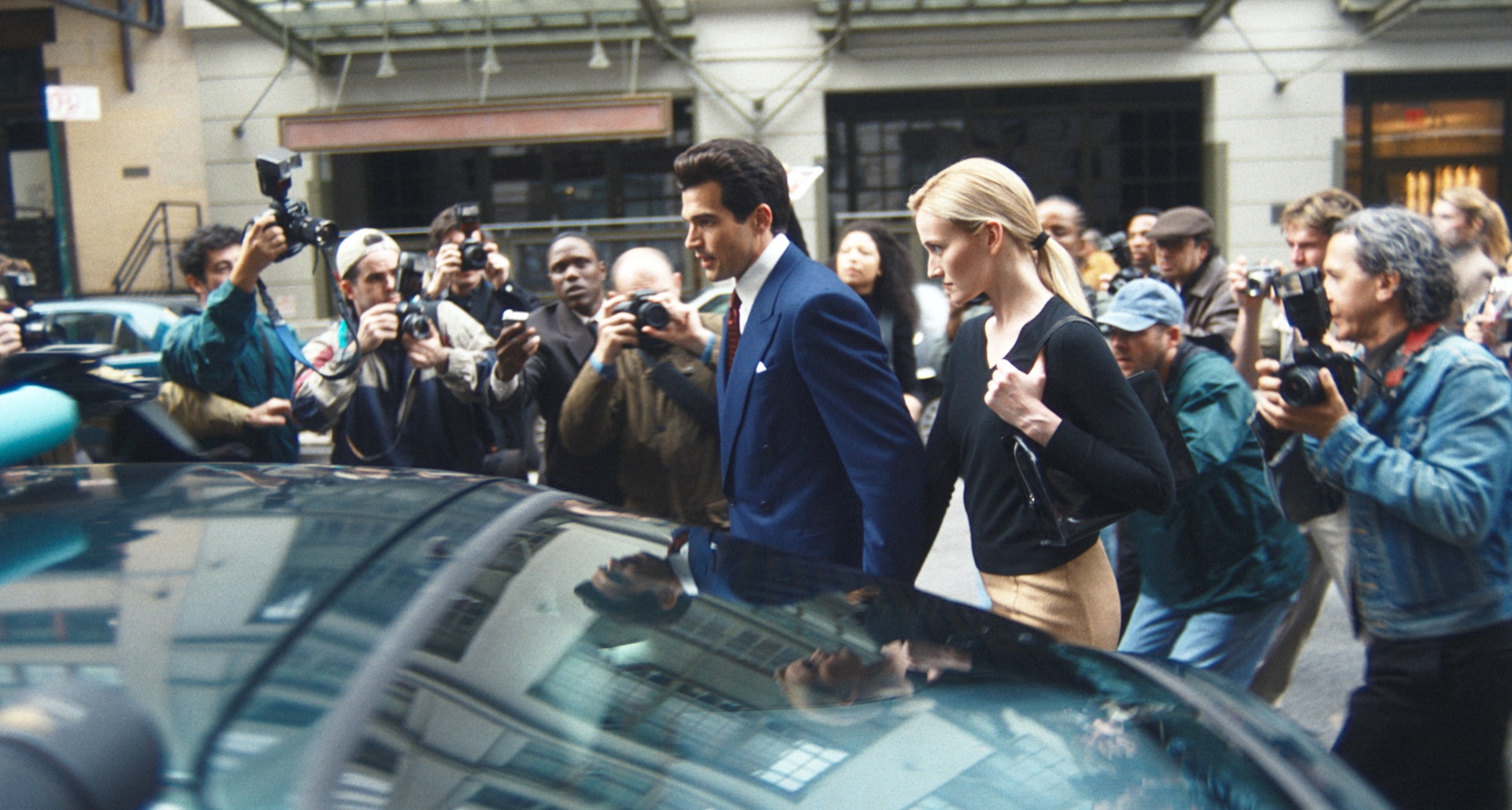 paul anthony kelly as john f kennedy jr and sarah pidgeon as carolyn bessette kennedy dodging paparazzi as they walk to a car in new york city in the tv show love story