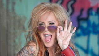 Lita Ford photographed in front of a grafitti'd wall holding her sunglasses above the bridge of her nose with her mouth open