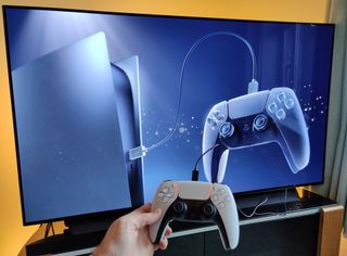 The PS5's DualSense controller being held by a hand in front of a TV.