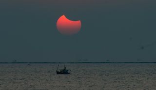 A partial solar eclipse shows a red sun with what looks like a moon shape "bite" take out of the top right corner. The sun sits in the sky above a body of water with a boat.