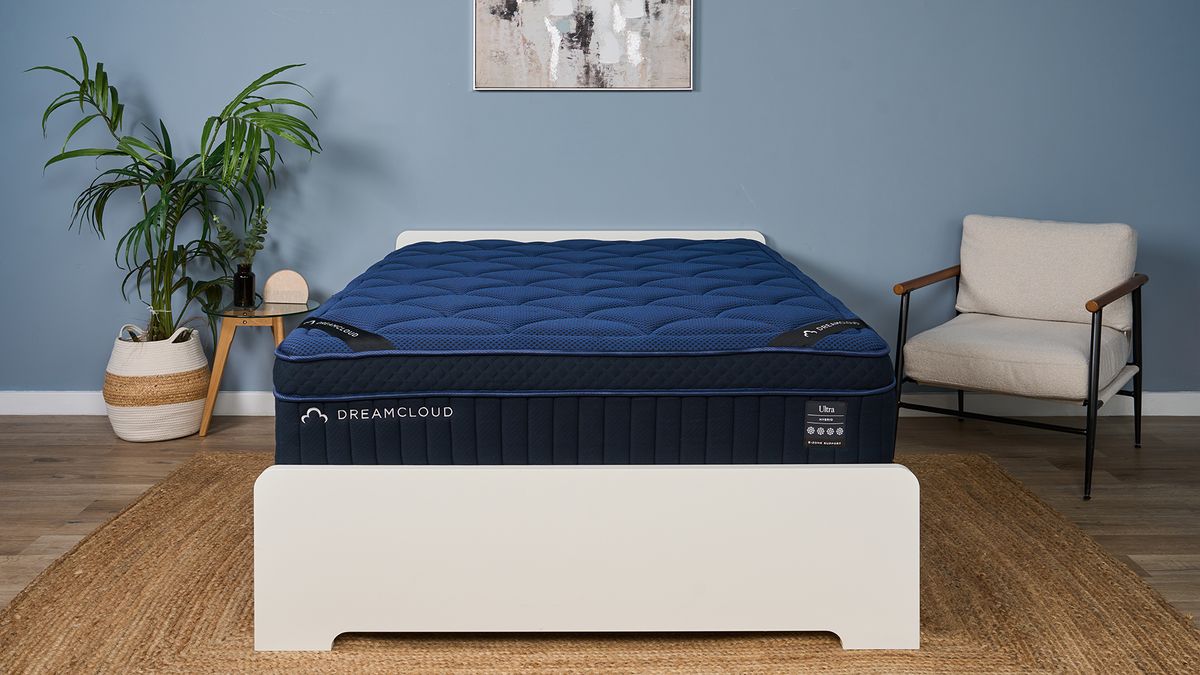 The image shows a navy blue mattress on a white bed frame, set within a muted blue bedroom