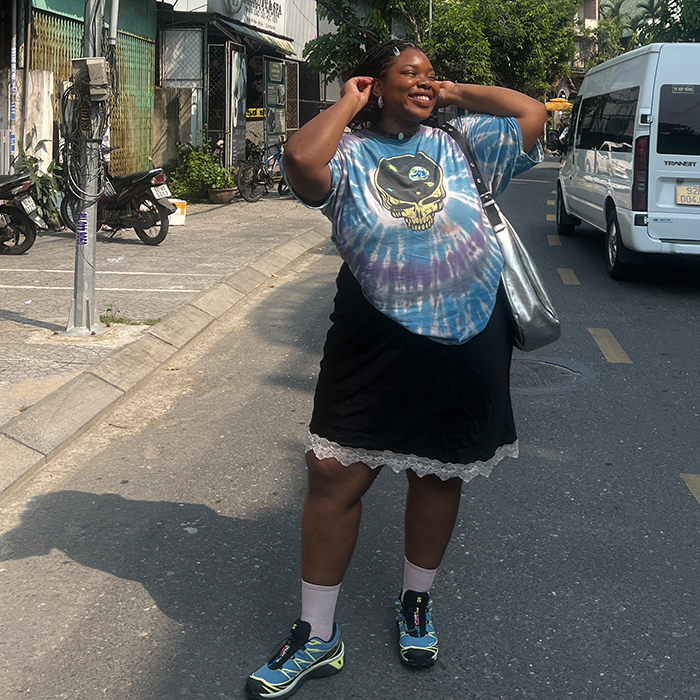 Chichi poses in the streets of Hoi An, Vietnam. She is wearing a blue tie-die graphic t-shirt, black skirt with lace trim, and blue Saloman sneakers while carrying a silver handbag. 