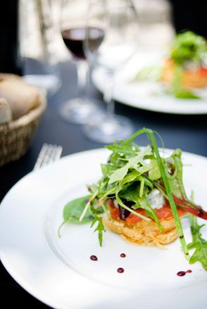 Close up of a white dinner plate with decorated food, blurred black table surface, drinking glasses one with half filled red wine, wicker basket with bread rolls