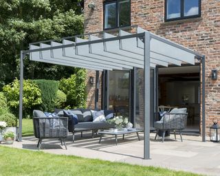 Architectural gray pergola above patio area, with grey garden furniture lounge set-up, on a paved patio next to grass